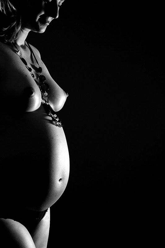 Die Schwangere guckt schmunzelnd auf ihren Bauch hinunter. Sie ist bis auf einen Slip und eine aus vielen Anhängern bestehenden Kette nackt.  Das Studiolicht des Fotografen Franz Fender aus Hannover modelliert ihre festen Brüste und ihren strammen Babybauch aus dem schwarzen Hintergrund heraus.