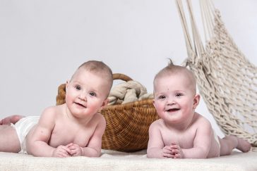 2 nur mit einer Windel bekleidete Babies liegen auf einem weißen Tuch. Sie liegen auf dem Bauch und stützen sich mit ihren Armen auf. Im Hintergrund steht ein Weidenkorb mit Tauen und eine Hängematte ragt angeschnitten ins Bild.